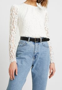 Hvid blonder bluse med lange ærmer, der har en rund halsudskæring og bølgede manchetter. Parret med højtaljede blå jeans og et sort bælte.