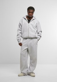 Felpa grigia con zip e pantaloni della tuta abbinati, realizzati in tessuto morbido, caratterizzati da una vestibilità comoda e un dettaglio del logo discreto. Indossati con scarpe di colore chiaro.