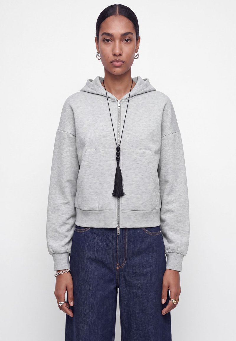 Modèle portant un sweat à capuche gris clair zippé, un jean foncé, des créoles, des bagues et un long collier noir à pompon, sur un fond uni.