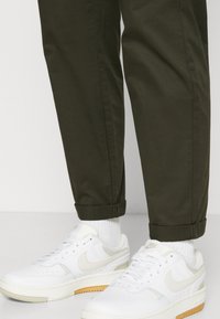 Des baskets Nike blanches avec des accents beige et une semelle en caoutchouc, associées à un pantalon olive roulé et des chaussettes blanches. Texture lisse.
