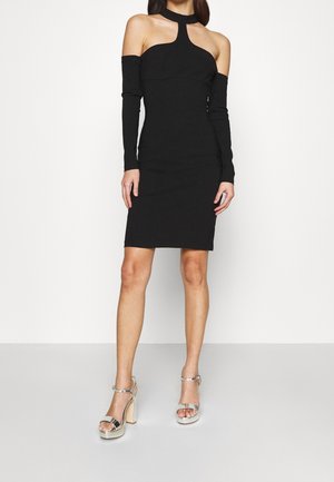 Czarna, dopasowana sukienka z długimi rękawami odsłaniającymi ramiona, dekoltem halter i długością do połowy uda. Połączona z srebrnymi sandałami na platformie.