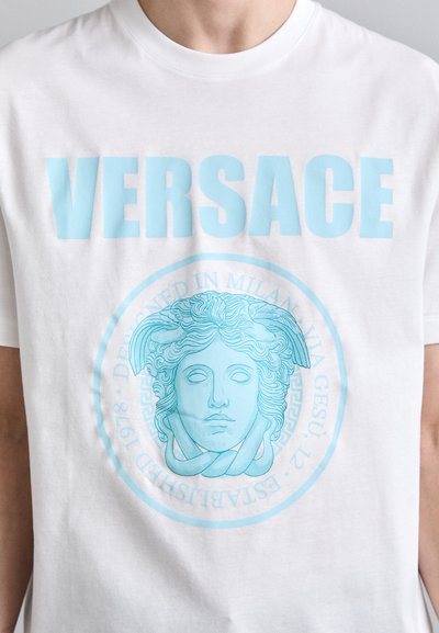 Balta kokvilnas T-krekls ar uzrakstu "VERSACE" gaisā zilā krāsā. Centrālā dizaina elementu veido zils medaljons ar klasisku seju.