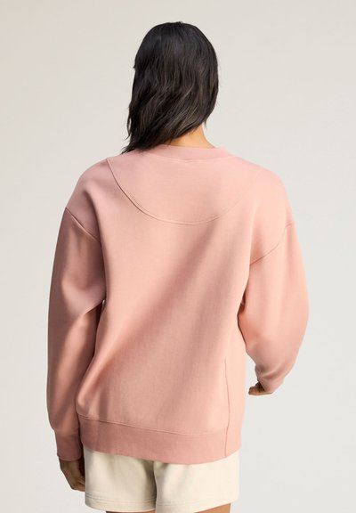 Person med mørkt hår iført en løs lyserød sweatshirt og beige shorts, vist bagfra mod en ensartet lys baggrund.