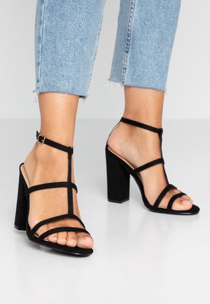 Fødder iført sorte høje hælsandaler med ankelremme kombineret med lyseblå cropped denimjeans på en hvid gulv.