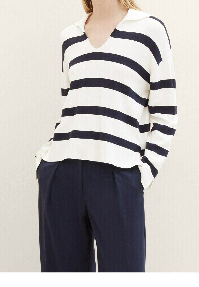 Femme portant un haut blanc à manches longues avec de larges rayures horizontales noires et un pantalon plissé bleu marine, les mains dans les poches.