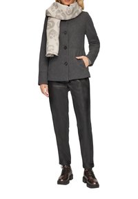 Manteau en laine mélange grise texturée avec deux boutons, poches latérales, et une écharpe beige avec un subtil motif animal. Pantalons en simili cuir noir et bottes marron.
