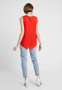 Czerwona top bez rękawów o asymetrycznym dołem, wykonany z lekkiego materiału, z subtelnym zapięciem na guziki z tyłu, zestawiony z jasnoniebieskimi dżinsami i białymi butami na słupku.