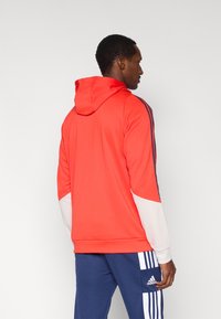 Sweat à capuche rouge avec accents blancs et bleus, doté de manches longues, d'une poche kangourou et de bandes sportives sur les côtés.