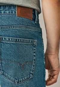 Jeans din denim albastru, cu un buzunar posterior cusut care are un detaliu unic sub formă de buclă. Pata din piele afișează "Bunuri de denim semnate".