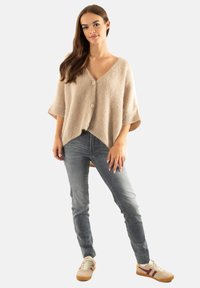 Cardigan beige avec un col en V, des manches courtes et deux boutons clairs. Associé à un jean gris et des baskets beiges avec des accents violets.