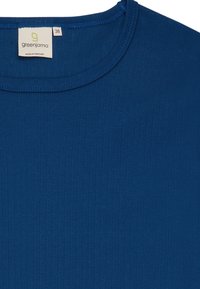 T-shirt blu a coste con colletto rotondo, realizzato in tessuto morbido. Presenta un'etichetta con "greenjama" e taglia 38, mostrando una texture liscia e un colore uniforme.