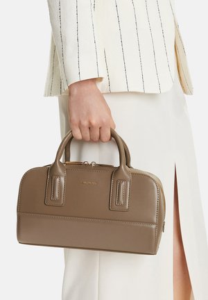 Mano che tiene una borsa strutturata in pelle taupe con manici superiori, indossata con pantaloni crema e una giacca bianca a righe nere.