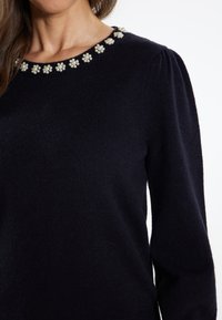 Maglione lavorato a maglia blu navy con scollatura rotonda decorata con elementi floreali simili a perle, maniche lunghe e una texture morbida.