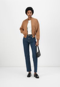 Chaqueta bomber marrón sobre una camisa blanca, combinada con jeans azul oscuro. Un bolso negro y mocasines completan el atuendo. Diseño y texturas sencillos.