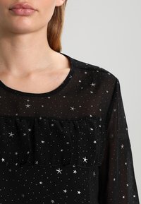 Chemisier noir transparent orné d'étoiles argentées, avec un col rond et un panneau froncé sur la poitrine. Design élégant et léger.