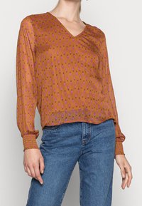 Braune, leichte Bluse mit langen Ärmeln und V-Ausschnitt, die mit einem rosa Blumenmuster verziert ist. Kombiniert mit hochtaillierten blauen Jeans.
