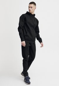 Svart hettegenser med frontlomme, justerbar snor og ribbestrikkede ermer, kombinert med matchende svarte joggebukser og mørke sneakers.