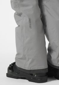Šedé lyžařské kalhoty s logem technologie RECCO®, mají zesílený lem, odolnou tkaninu a jsou doplněny černými snowboardovými botami.