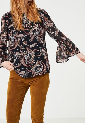 Kvinde iført en sort paisley-mønstret bluse med vide ærmer og sennepsfarvede kordfløjlsbukser mod en ensfarvet baggrund.