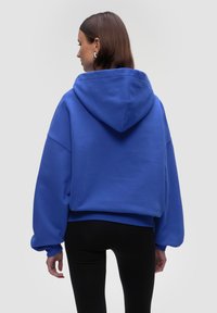 Felpa blu con cappuccio, dalla vestibilità rilassata, cappuccio grande e polsini a coste, abbinata a leggings neri, mostrata di spalle.