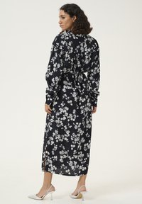 Robe maxi noire à motifs floraux avec des manches longues, une taille nouée et des fentes sur les côtés. Présente des motifs floraux blancs et une texture de tissu lisse et fluide.