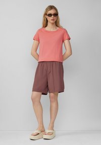 Koralfarbenes T-Shirt mit kurzen Ärmeln, kombiniert mit braunen Shorts. Das Model trägt Plateausandalen. Schlichtes Design mit elastischem Bund an den Shorts.