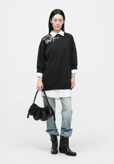 Μαύρο oversized φούτερ με floral διακοσμήσεις, πάνω από λευκό πουκάμισο, συνδυασμένο με ανοιχτό μπλε τζιν και μαύρες μπότες μέχρι τον αστράγαλο, κρατώντας μια μαύρη τσάντα.