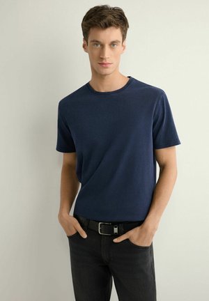 Młody mężczyzna z krótkimi brązowymi włosami, ubrany w granatowy krótki rękaw t-shirt oraz czarne jeansy, z rękami w kieszeniach, stoi na jednolitym tle.