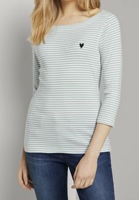 Frau in einem langärmeligen weißen Hemd mit hellgrünen Streifen und einem kleinen schwarzen Herz auf der Brust, kombiniert mit blauen Jeans.