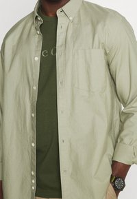 Lys grøn knap-up skjorte over en olivengrøn t-shirt, med en brystlomme og et tekstureret stof. Outfittet komplementeres af et mørkt ur.