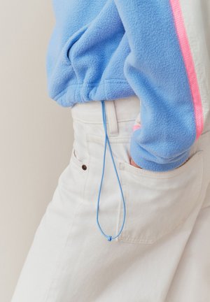 Persona que lleva pantalones blancos con la mano en el bolsillo trasero y una chaqueta de forro polar azul con una raya rosa y cordón azul suelto.