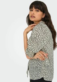 Lichtgekleurde blouse met zwarte abstracte vlekken, lange mouwen, knoopsluiting aan de voorkant en strikdetail in de taille. Gladde stofstructuur.