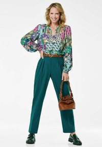 Blusa multicolore a fantasia con maniche a sbuffo, pantaloni teal a vita alta, cintura marrone e una piccola borsa marrone. Scarpe platform verdi.