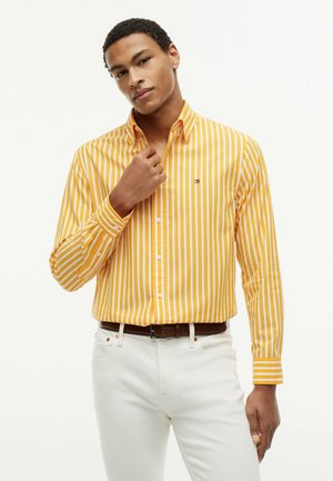 Mann richtet den Kragen eines langärmeligen gelb-weiß gestreiften Hemdes, trägt weiße Hose und einen braunen Gürtel, steht vor weißem Hintergrund.