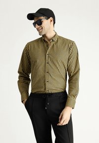Camisa de manga larga a rayas amarillas y negras con cuello abotonado, combinada con pantalones negros y una gorra negra. Presenta un corte relajado.