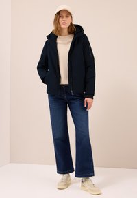 Giacca isolata blu navy con cappuccio, cerniera frontale e tasche laterali, indossata sopra un maglione di lana beige e jeans a gamba larga blu.