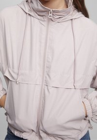 Light pink windbreaker with a hood, elastic cuffs, and a front zipper. Features a double-layered chest pocket and a smooth, matte texture.