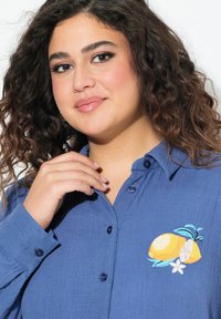 Camicia azzurra con bottoni realizzata in un tessuto testurizzato, decorata con un ricamo di limone, foglie verdi e un fiore bianco come accentuazione.
