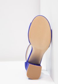 Sole modré otevřené sandály s blokovým podpatkem, béžovou texturovanou podrážkou a označením velikosti 37, umístěná proti bílé rohové zdi.