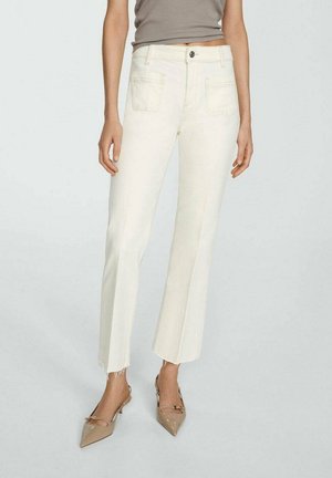 Kobieta ubrana w kremowe jeansy o prostych nogawkach z surowymi wykończeniami, beżowe szpilki z noskiem w szpic oraz bezrękawnik w kolorze taupe na jednolitym tle.