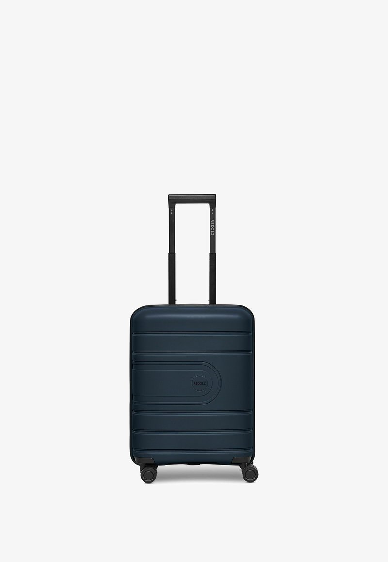 Harde koffer in diep marineblauw, met horizontale groeven, een centraal logo, uitschuifbare zwarte handgreep en vier zwarte wielen voor mobiliteit.