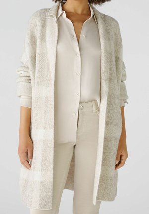 Femme portant un long cardigan à carreaux beige clair, un chemisier crème à boutons et un pantalon clair sur un fond neutre.