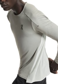 Maglietta sportiva a maniche lunghe di colore grigio chiaro con tessuto strutturato, scollo rotondo e logo nero sul petto. Caratterizzata da un design minimale.