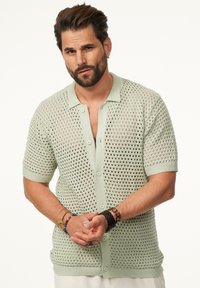Hellgrüner, kurzärmeliger Strickcardigan mit offenem Netzdesign, Knopfverschluss sowie geripptem Kragen und Bündchen.