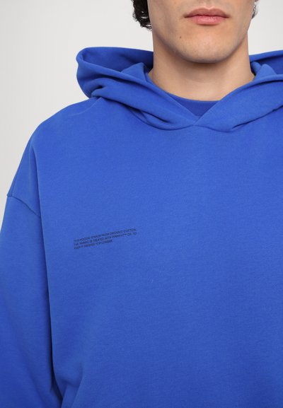 Mėlynas organinės medvilnės hoody, šiek tiek per didelio modelio, su priekinėmis kišenėmis ir spausdintu tekstu ant kairės krūtinės.