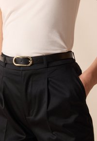 Czarny skórzany pasek z złotą owalną klamrą, noszony z wysokim stanem czarnych spodni i dopasowanym białym topem na ramiączkach, podkreślającym szyte plisy.