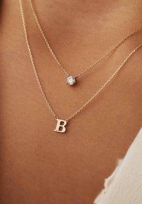 Goldene Halsketten mit einem geschichteten Design. Eine hat einen kreisförmigen klaren Steinanhänger, während die andere einen Anhänger in Form eines großen "B" hat.