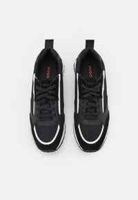 Chaussures de sport noires avec des matériaux en maille et en daim. Elles présentent des accents blancs, un système de laçage et une boucle pour les enfiler facilement. Marque visible sur la semelle intérieure.