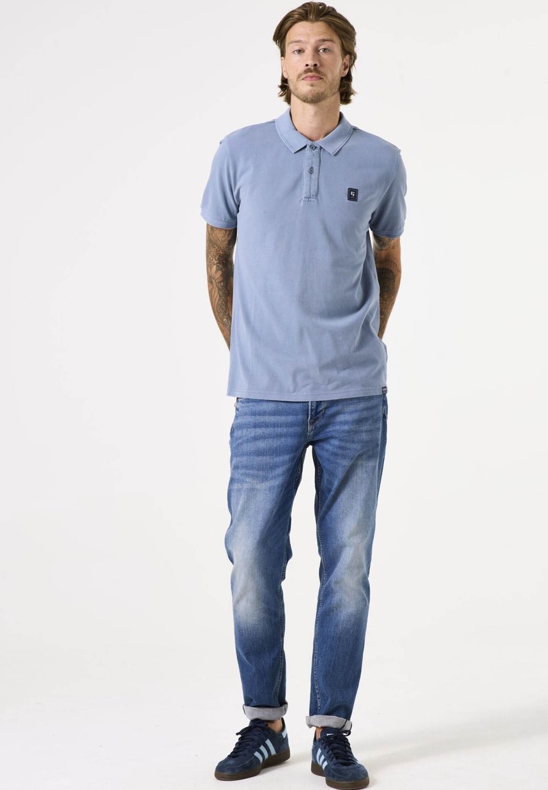 Chemise polo bleu clair avec un col et trois boutons, associée à un jean bleu délavé et des baskets navy avec des rayures bleu clair.