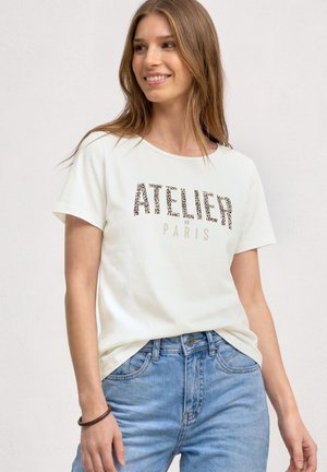 Junge Frau mit langen hellbraunen Haaren, die ein weißes T-Shirt mit dem Text "ATELIER DE PARIS" und blaue Jeans trägt, lächelt und schaut zur Seite.